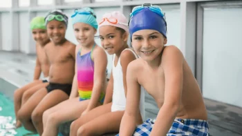 Kids sitting on the edge of a pool in swimsuits, smiling.