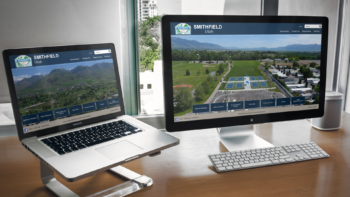 A desktop computer and a laptop on a stand, both displaying the website homepage of the City of Smithfield, UT